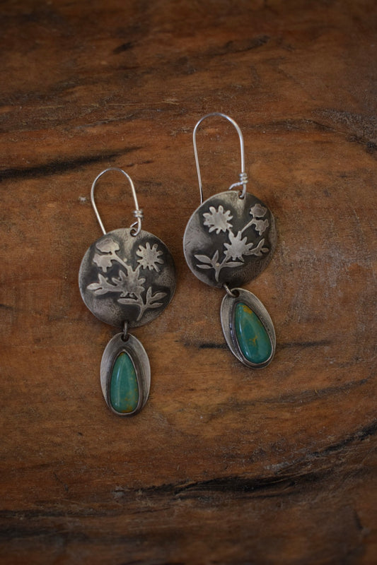 The Wildflower earrings have a domed round silver backplate with flowers on the front. Below hang two green teardrop shaped turquoise.
