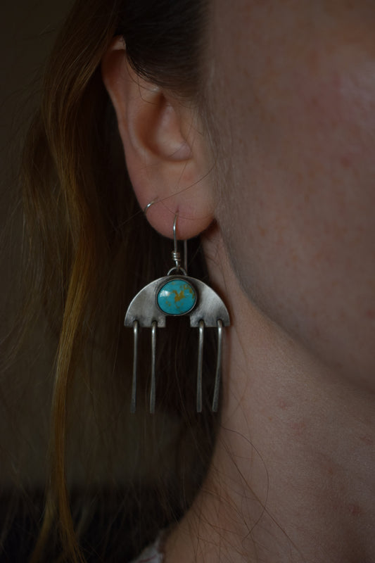 A round blue turquoise sits on a half round cutout silver backplate. Four silver wires hang below.