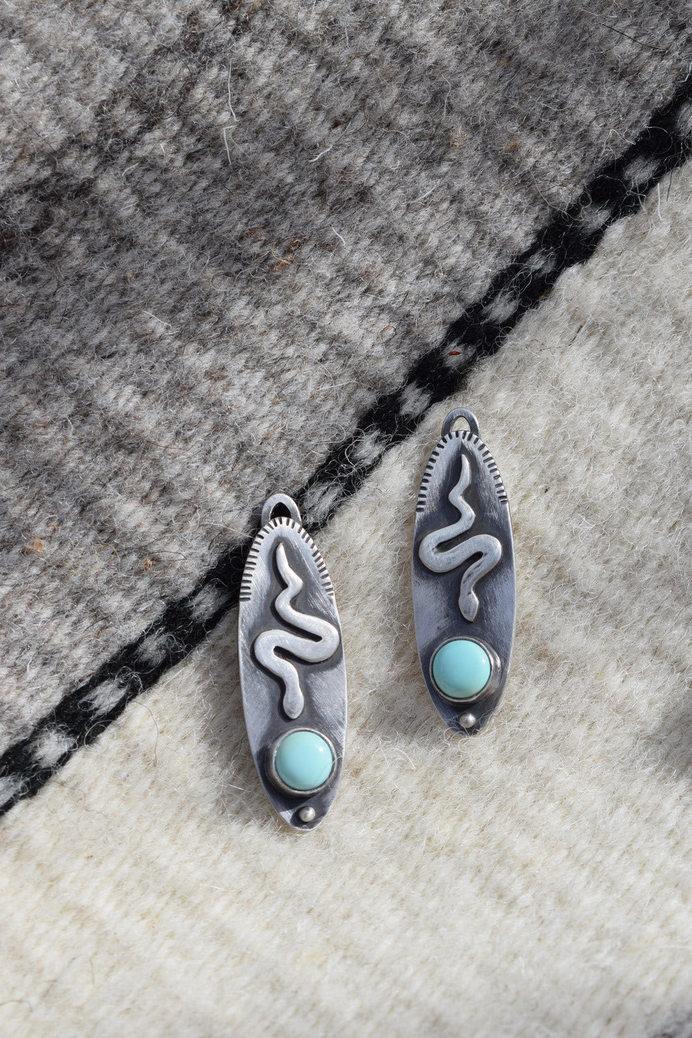The Serpent Sister earrings are a long oval backplate with cutout snakes at the top. They face small green dots of turquoise at the bottom. The sides are line stamped.